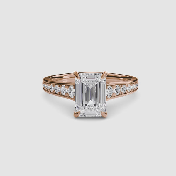 Rose gold ring with a large emerald cut diamond and smaller round diamonds on the band against a white background