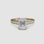 Gold ring with a large emerald cut diamond and smaller round diamonds on the band against a white background