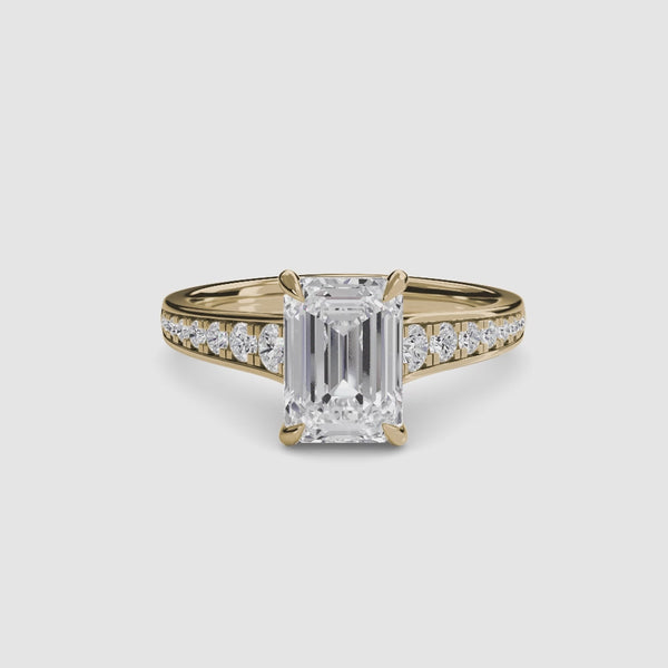 Gold ring with a large emerald cut diamond and smaller round diamonds on the band against a white background