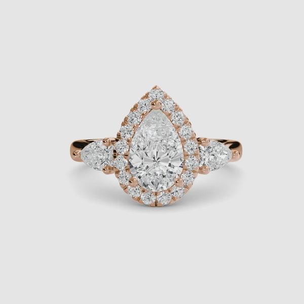 Rose gold three stone ring with a pear-shaped diamond and halo on a white background