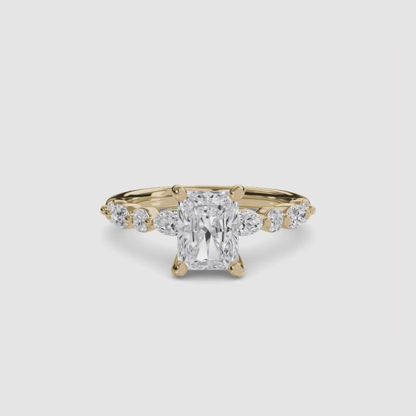 Gold ring with a central radiant cut diamond and marquise and round side stones on the band on a white background